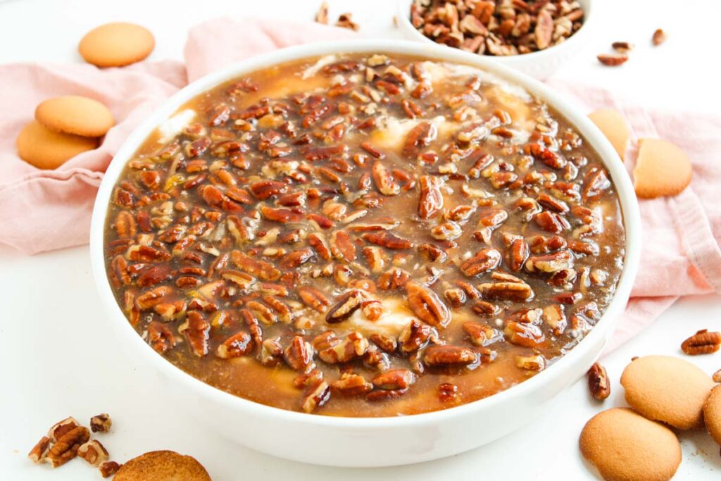 A round white dish filled with Pecan Pie Dip, topped with pecans, possibly resembling pudding or cobbler, surrounded by pecans and vanilla wafer cookies on a white surface.