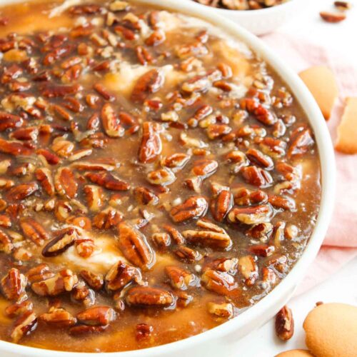 A close-up of Pecan Pie Dip topped with glazed pecans, with a bowl of extra pecans and vanilla wafer cookies nearby on a light surface.