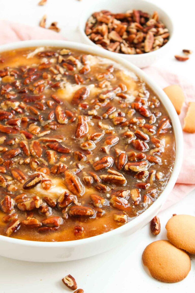 A close-up of Pecan Pie Dip topped with glazed pecans, with a bowl of extra pecans and vanilla wafer cookies nearby on a light surface.