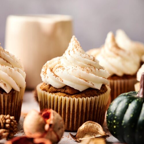 Close-up of Pumpkin Cupcakes with swirled cream frosting, surrounded by autumnal decorations and a ceramic mug in the background.