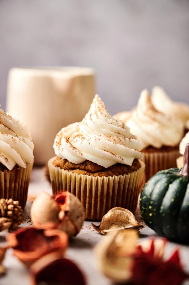 Close-up of Pumpkin Cupcakes with swirled cream frosting, surrounded by autumnal decorations and a ceramic mug in the background.
