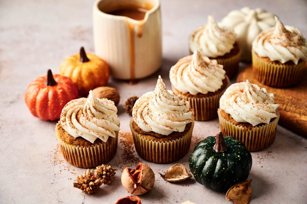 Pumpkin cupcakes with white frosting are arranged on a table with decorative pumpkins, walnuts, and a small pitcher of caramel sauce in the background.