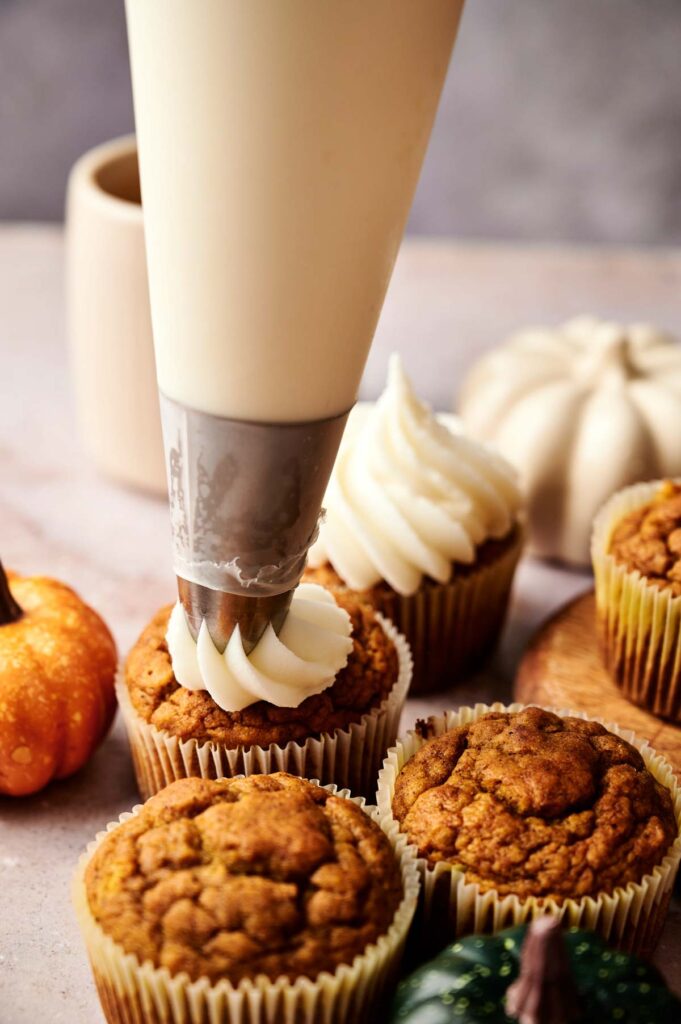 A piping bag is used to frost pumpkin cupcakes on a table, surrounded by several unfrosted cupcakes and small decorative pumpkins nearby.
