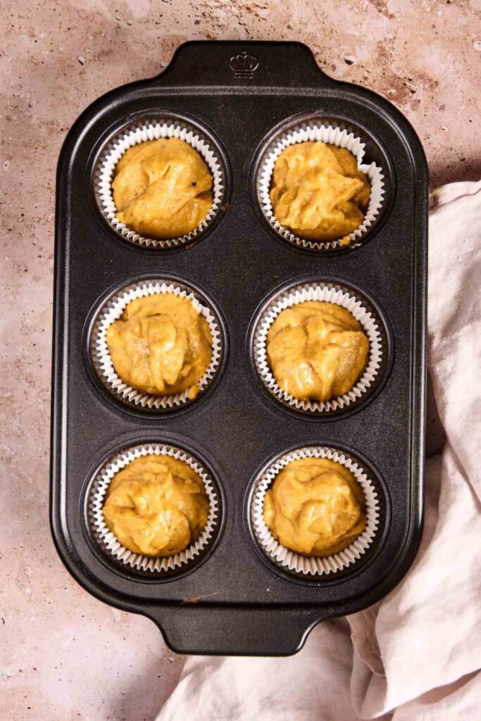 A muffin tin holds six paper liners filled with unbaked pumpkin cupcakes batter, placed on a light countertop next to a beige cloth.