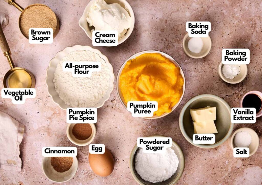 Top-down view of labeled baking ingredients for Pumpkin Cupcakes, including brown sugar, cream cheese, baking soda, baking powder, flour, pumpkin puree, butter, powdered sugar, vanilla, egg, spices, salt, and oil.