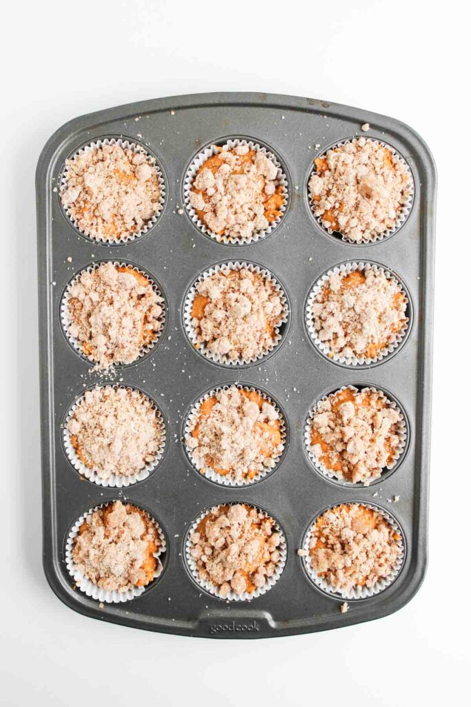 A muffin tin filled with twelve paper-lined cups, each containing unbaked Pumpkin Spice Muffins batter topped with a crumbly streusel—perfect when finished with a swirl of cream cheese frosting.