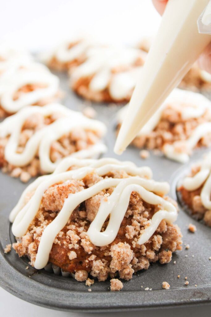 A hand decorates Pumpkin Spice Muffins with Cream Cheese Frosting, zigzagging white icing over a crumbly topping in a muffin tin.