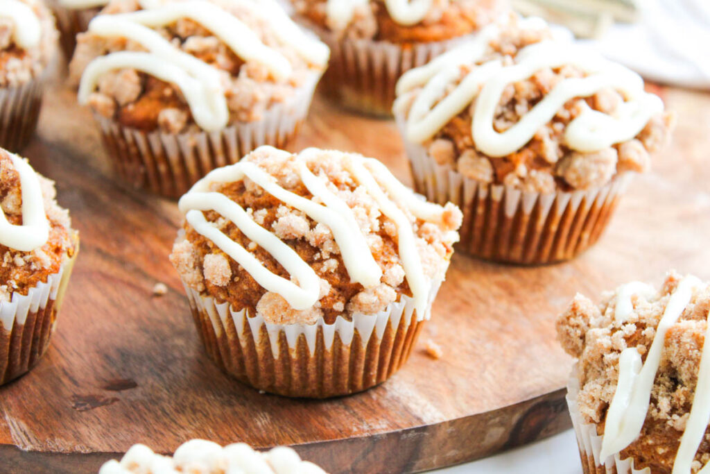 Pumpkin Spice Muffins with Cream Cheese Frosting crumb topping and white icing drizzle are arranged beautifully on a wooden serving board.