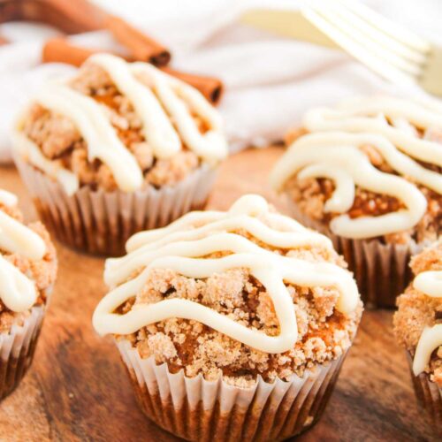 Five Pumpkin Spice Muffins with Cream Cheese Frosting are crumb-topped and drizzled with white icing, arranged on a wooden surface with a gold fork and cinnamon sticks in the background.