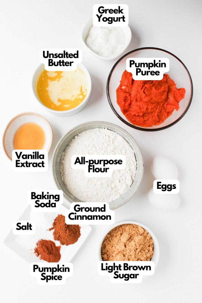 Top-down view of labeled baking ingredients for Pumpkin Spice Muffins with Cream Cheese Frosting, including pumpkin puree, flour, Greek yogurt, eggs, butter, vanilla extract, sugar, spices, salt, and baking soda.