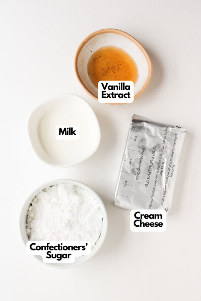 Four labeled ingredients for frosting on a white surface: vanilla extract, milk, cream cheese in foil, and confectioners' sugar in a bowl—perfect for topping Pumpkin Spice Muffins with Cream Cheese Frosting.