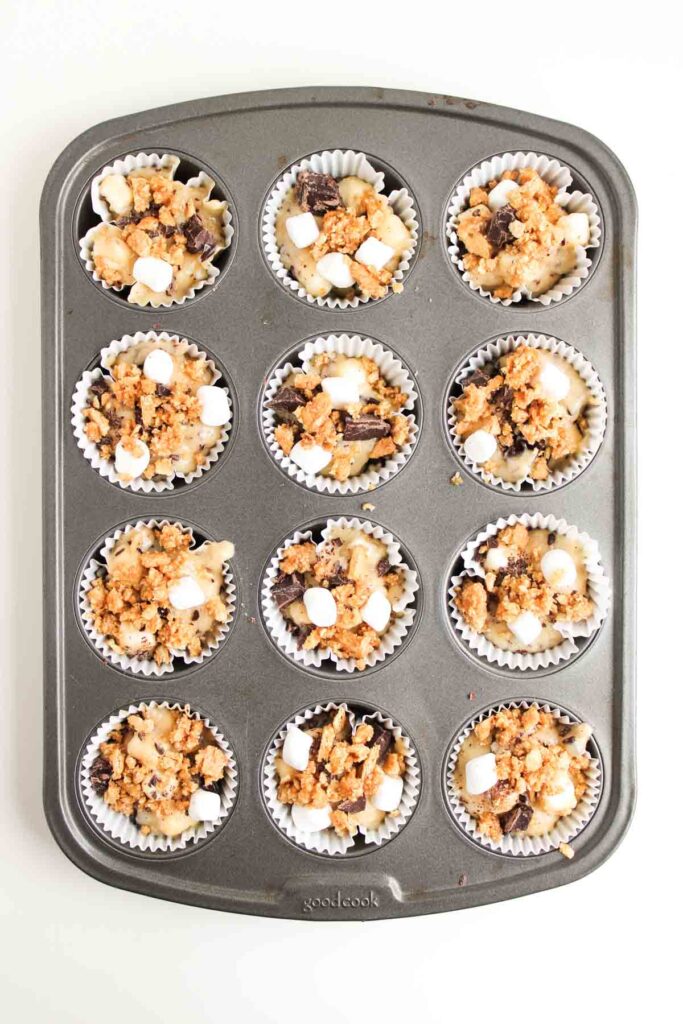 A muffin tin with twelve paper liners, each filled with a mixture of pasta, mini marshmallows, chocolate chunks, and crumb topping—these inventive S'mores Muffins are ready to be baked.