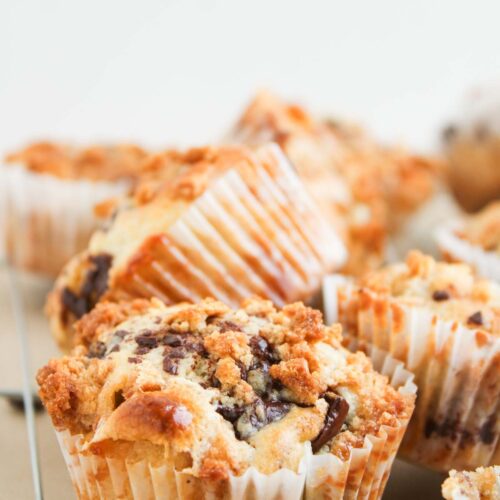 S'mores Muffins with chocolate chips and crumb topping cool on a wire rack in white paper liners, set against a plain background.