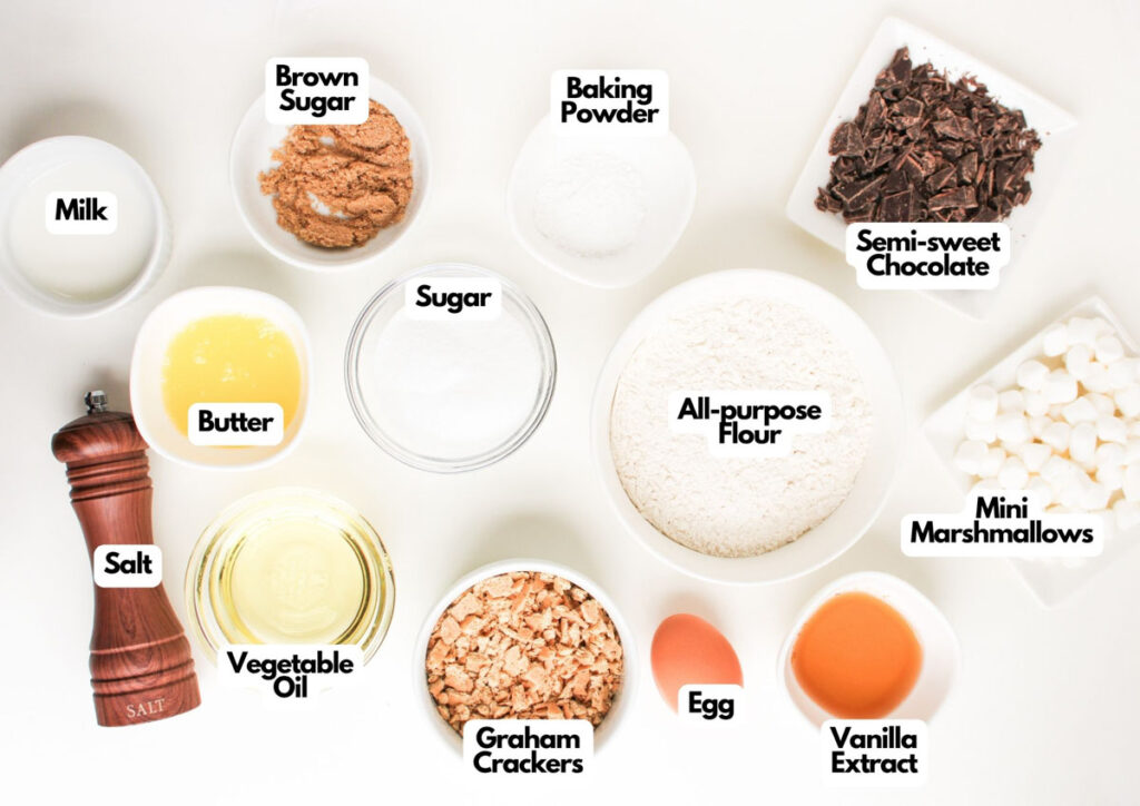 Top-down view of labeled bowls and containers holding ingredients for S'mores Muffins, including milk, brown sugar, baking powder, semi-sweet chocolate, sugar, butter, vegetable oil, salt, graham crackers, flour, egg, vanilla, and mini marshmallows.