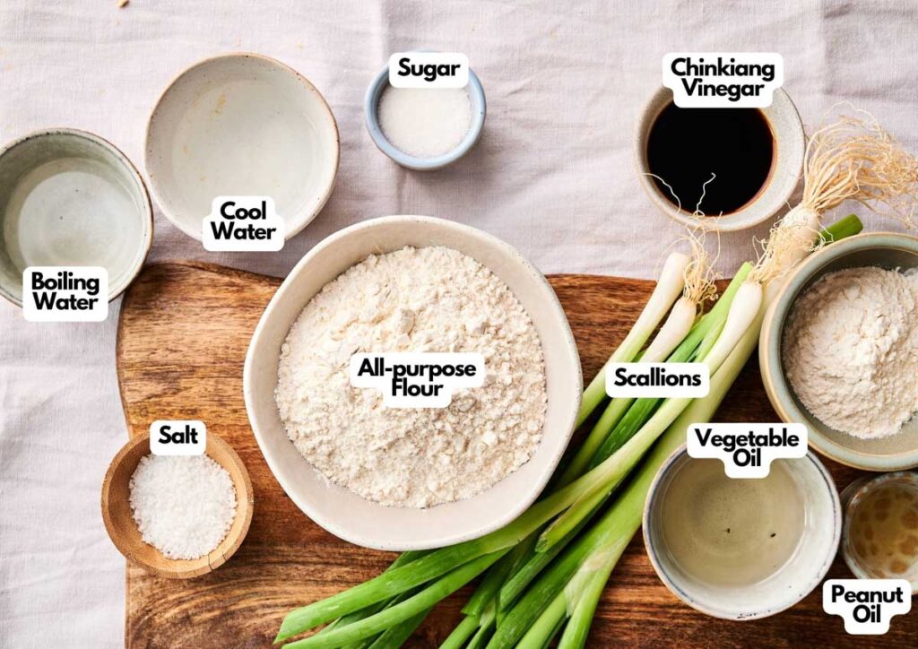A wooden surface displays ingredients for Scallion Pancakes&mdash;bowls of all-purpose flour, boiling water, cool water, salt, sugar, Chinkiang vinegar, peanut oil, vegetable oil, and fresh scallions.