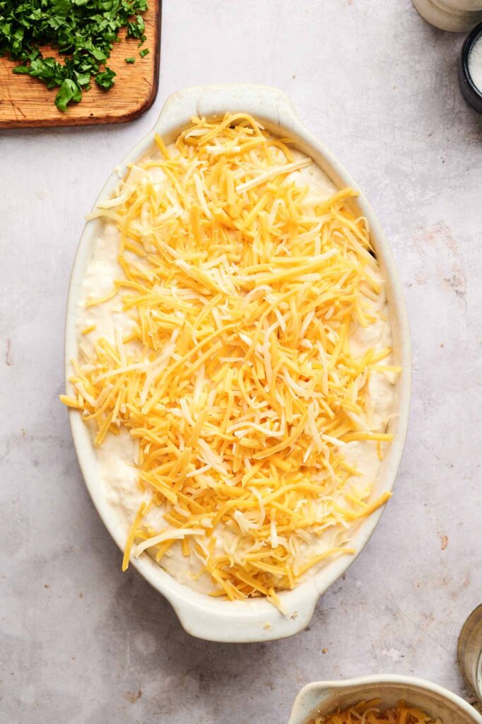 An oval baking dish filled with a creamy Sloppy Joe Casserole mixture is topped with shredded yellow and white cheese, ready for baking. Chopped herbs are on a cutting board nearby.