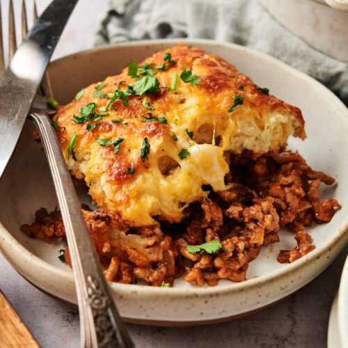 A serving of shepherd's pie with a golden, cheesy mashed potato topping and seasoned ground meat filling, reminiscent of a Sloppy Joe Casserole, garnished with chopped parsley on a ceramic plate with a fork and knife.