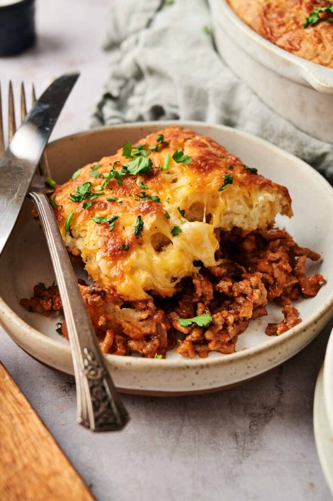 A serving of shepherd's pie with a golden, cheesy mashed potato topping and seasoned ground meat filling, reminiscent of a Sloppy Joe Casserole, garnished with chopped parsley on a ceramic plate with a fork and knife.
