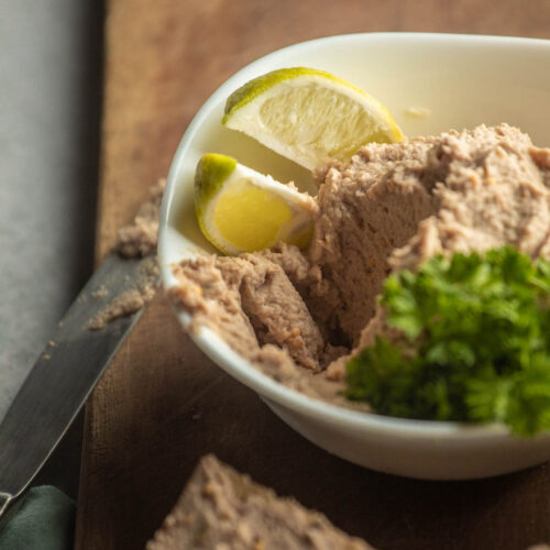 A white bowl filled with smoked mackerel pate, garnished with lime wedges and parsley, sits on a wooden board next to a knife.