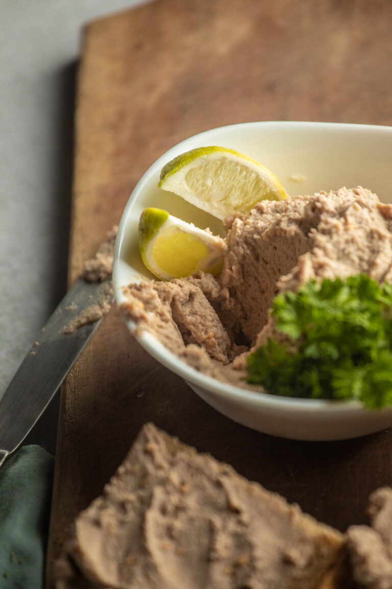 A white bowl filled with smoked mackerel pate, garnished with lime wedges and parsley, sits on a wooden board next to a knife.