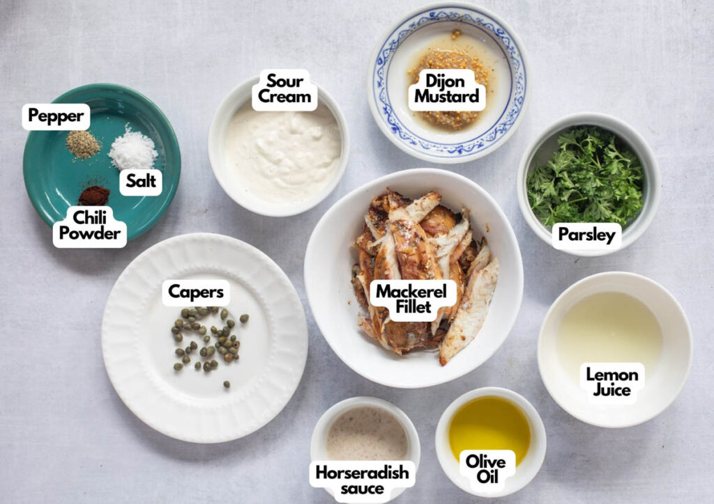 A flat lay of labeled bowls containing ingredients for Smoked Mackerel Pate: mackerel fillet, capers, sour cream, Dijon mustard, parsley, lemon juice, olive oil, horseradish sauce, chili powder, salt, and pepper.