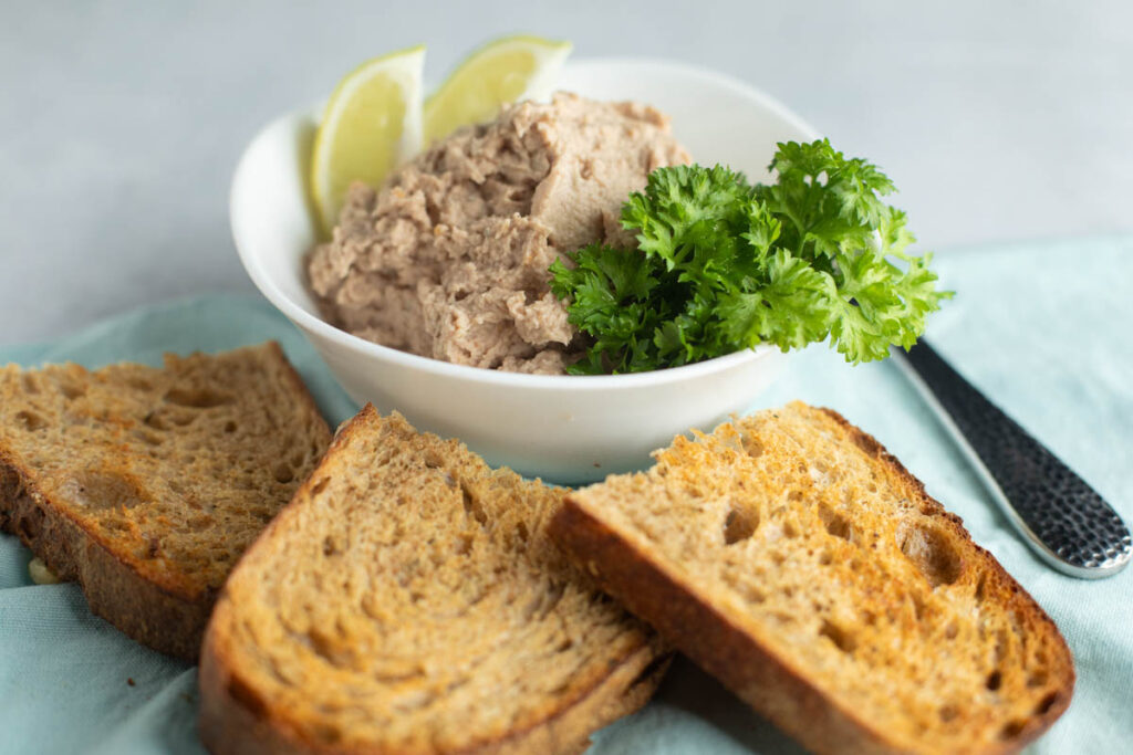 A bowl of Smoked Mackerel Pate garnished with parsley and lime wedges, served with three slices of toasted bread on a blue cloth.