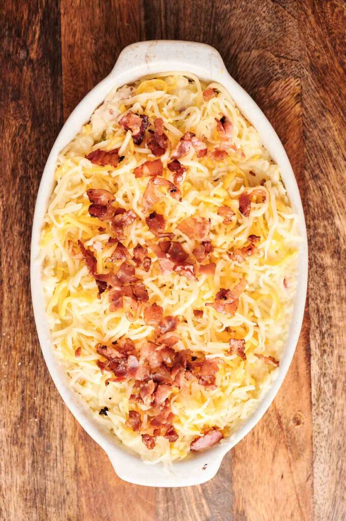 An oval baking dish filled with shredded cheese and cooked bacon pieces on top, this Twice-Baked Potato Casserole sits invitingly on a wooden surface.