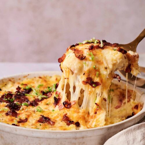 A serving of cheesy Twice-Baked Potato Casserole topped with crispy bacon and chopped green onions is being lifted from a round baking dish.