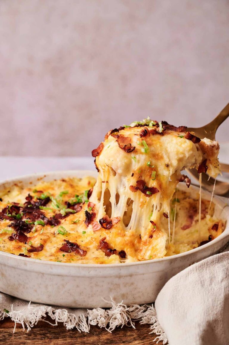A serving of cheesy Twice-Baked Potato Casserole topped with crispy bacon and chopped green onions is being lifted from a round baking dish.