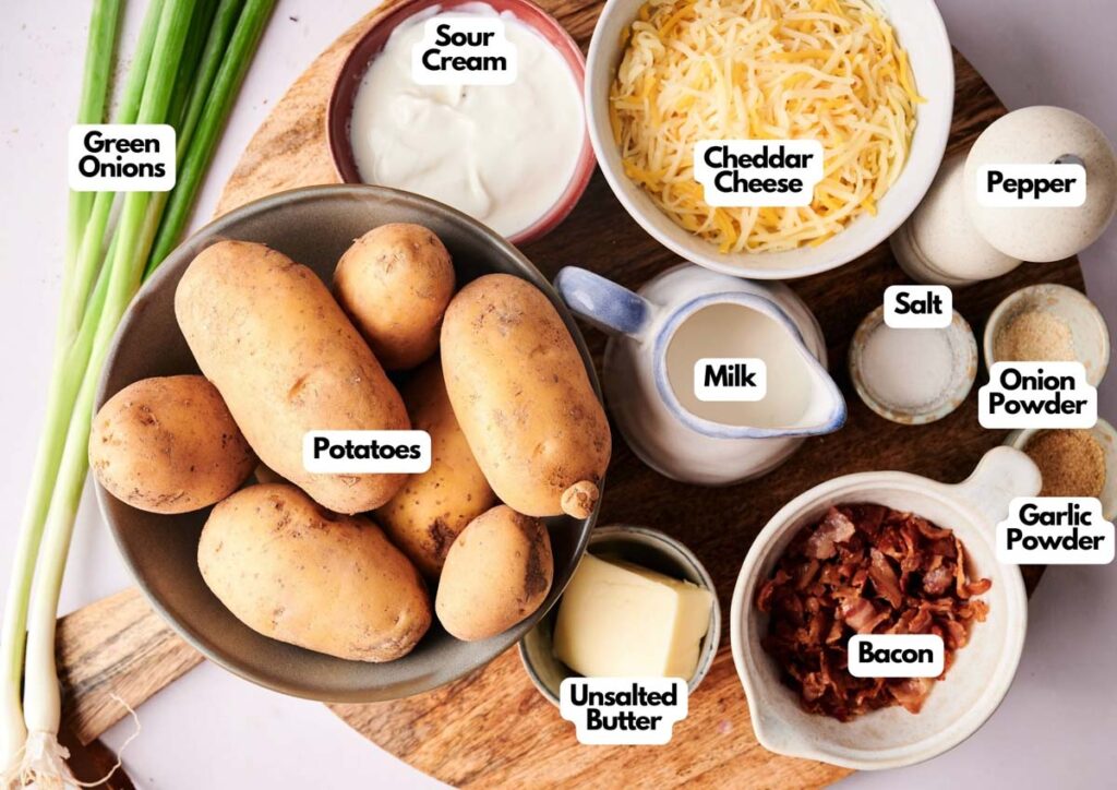 A tabletop displays ingredients for Twice-Baked Potato Casserole: potatoes, green onions, sour cream, cheddar cheese, milk, unsalted butter, bacon, salt, pepper, onion powder, and garlic powder in separate bowls and containers.