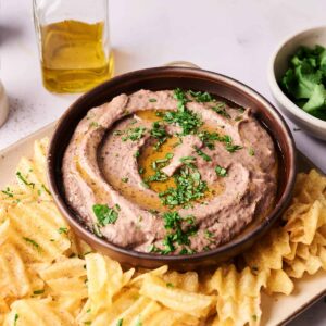 A bowl of Black Bean Hummus garnished with chopped herbs and olive oil, surrounded by waffle-cut potato chips on a rectangular plate. A bottle of olive oil and a bowl of herbs are visible in the background.