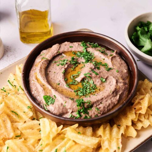 A bowl of Black Bean Hummus garnished with chopped herbs and olive oil, surrounded by waffle-cut potato chips on a rectangular plate. A bottle of olive oil and a bowl of herbs are visible in the background.