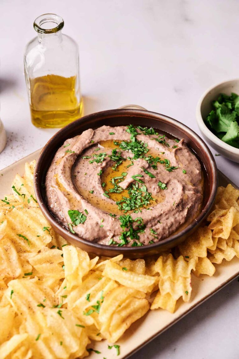 A bowl of Black Bean Hummus garnished with chopped herbs and olive oil, surrounded by waffle-cut potato chips on a rectangular plate. A bottle of olive oil and a bowl of herbs are visible in the background.