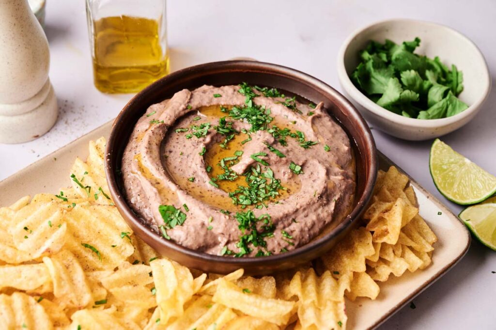 A bowl of Black Bean Hummus dip garnished with chopped herbs and olive oil, surrounded by waffle potato chips, with lime wedges and cilantro on the side.