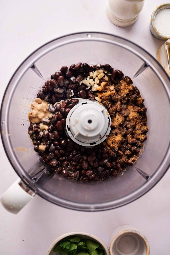 Top view of a food processor filled with black beans, spices, garlic, and more&mdash;ready to blend into Black Bean Hummus. Small bowls with extra ingredients are arranged nearby for easy access.