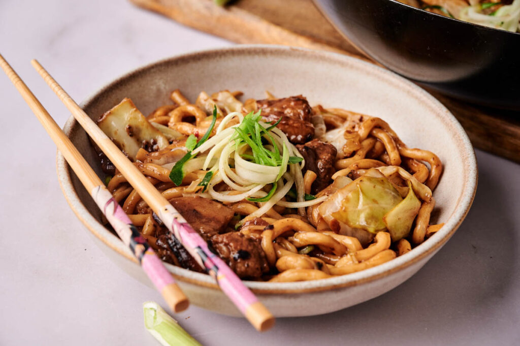 A bowl of stir-fried black bean noodles with beef, cabbage, and green onion garnish, served with chopsticks resting on the bowl&rsquo;s edge.