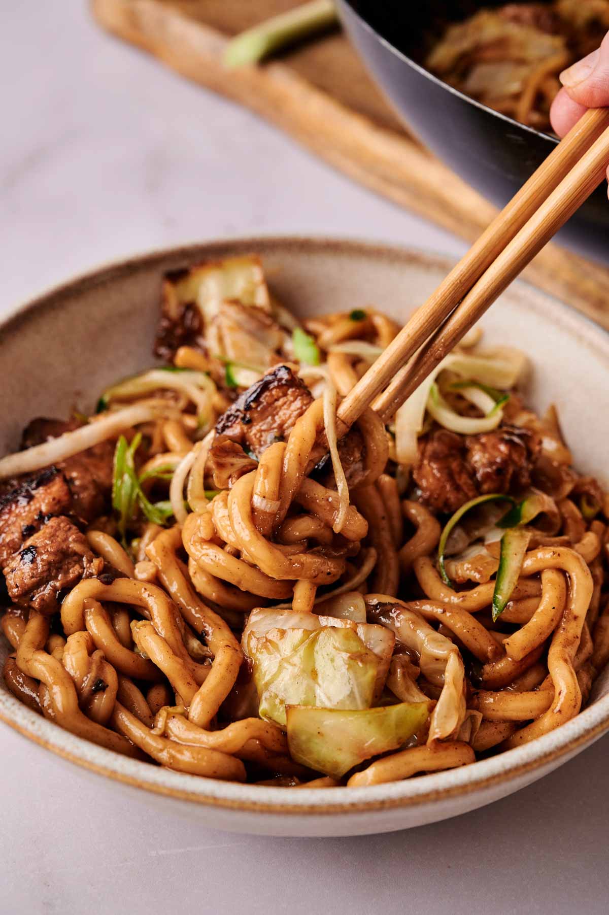 A bowl of stir-fried udon noodles with vegetables and pieces of tofu, inspired by Black Bean Noodles, with chopsticks picking up a portion.