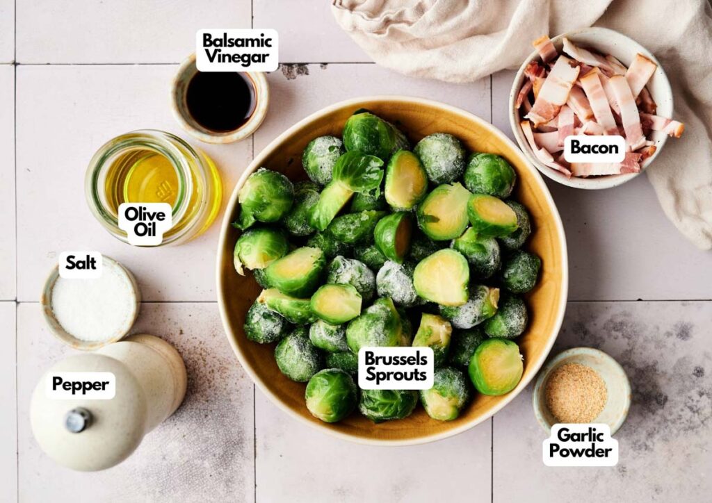 A bowl of Brussels Sprouts with Bacon sits on a tiled surface, surrounded by labeled ingredients: garlic powder, balsamic vinegar, olive oil, salt, and pepper.
