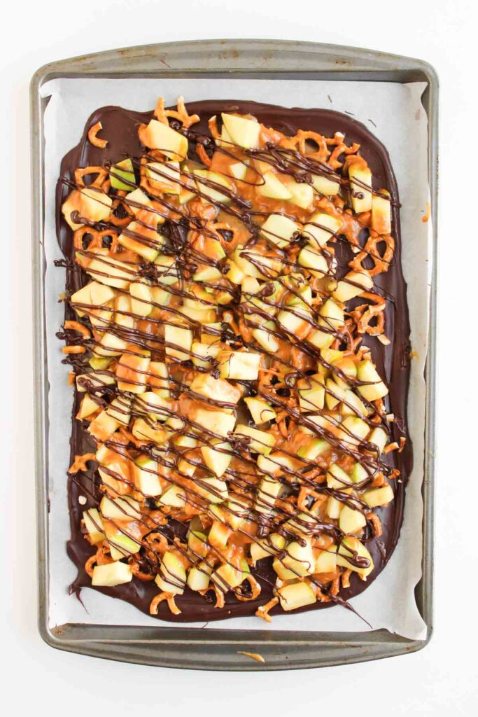 A baking sheet with Caramel Apple Bark, topped with chopped apples, pretzels, caramel drizzle, and dark chocolate on parchment paper.