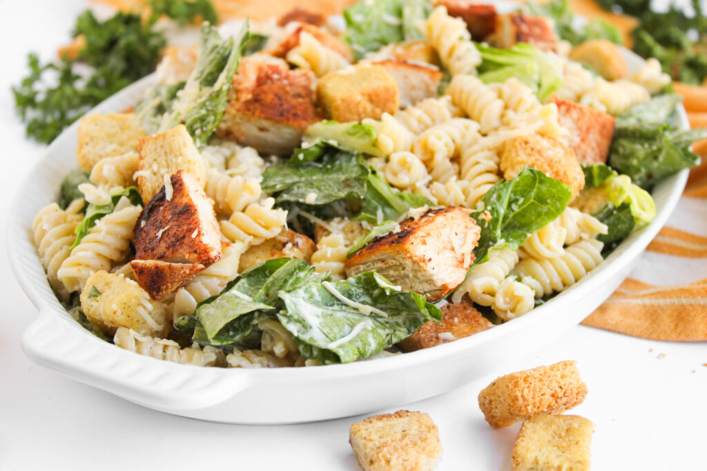 A white dish filled with Chicken Caesar Pasta Salad—rotini pasta, grilled chicken pieces, romaine lettuce, croutons, and creamy dressing.
