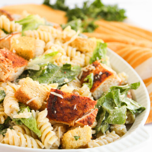 A plate of Chicken Caesar Pasta Salad with rotini, romaine lettuce, croutons, shredded cheese, and creamy dressing, served alongside a baguette and parsley in the background.