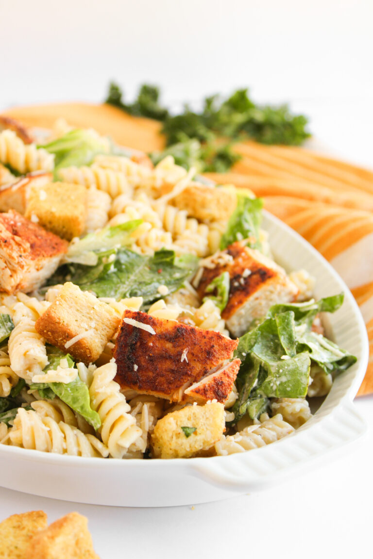 A plate of Chicken Caesar Pasta Salad with rotini, romaine lettuce, croutons, shredded cheese, and creamy dressing, served alongside a baguette and parsley in the background.