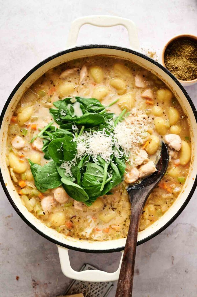 A pot of creamy Chicken Gnocchi Soup with tender chicken and vegetables, topped with fresh spinach and grated cheese, with a wooden spoon resting inside.