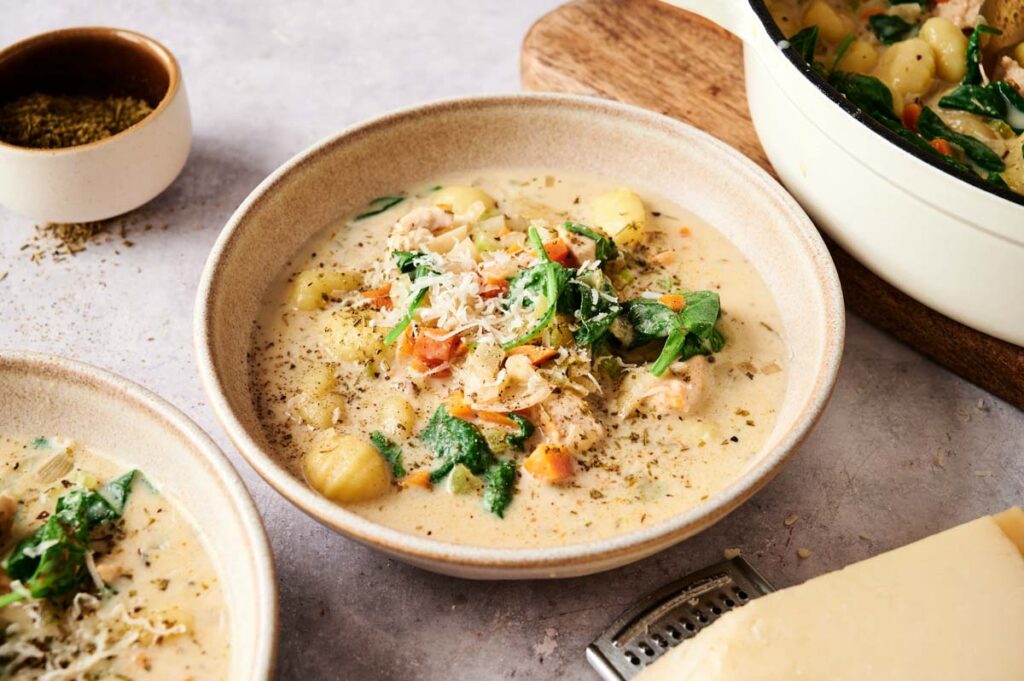 A bowl of creamy Chicken Gnocchi Soup with spinach, chicken, and carrots, topped with grated cheese and herbs. In the background, a pot of soup and fresh ingredients complete the cozy scene.