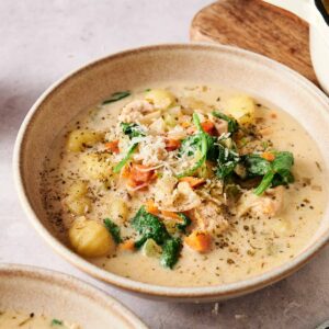 A bowl of creamy Chicken Gnocchi Soup, garnished with grated cheese and herbs, sits on a table next to a pot and a wooden board.