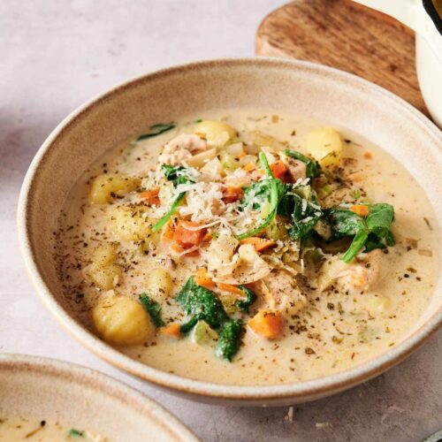 A bowl of creamy Chicken Gnocchi Soup, garnished with grated cheese and herbs, sits on a table next to a pot and a wooden board.