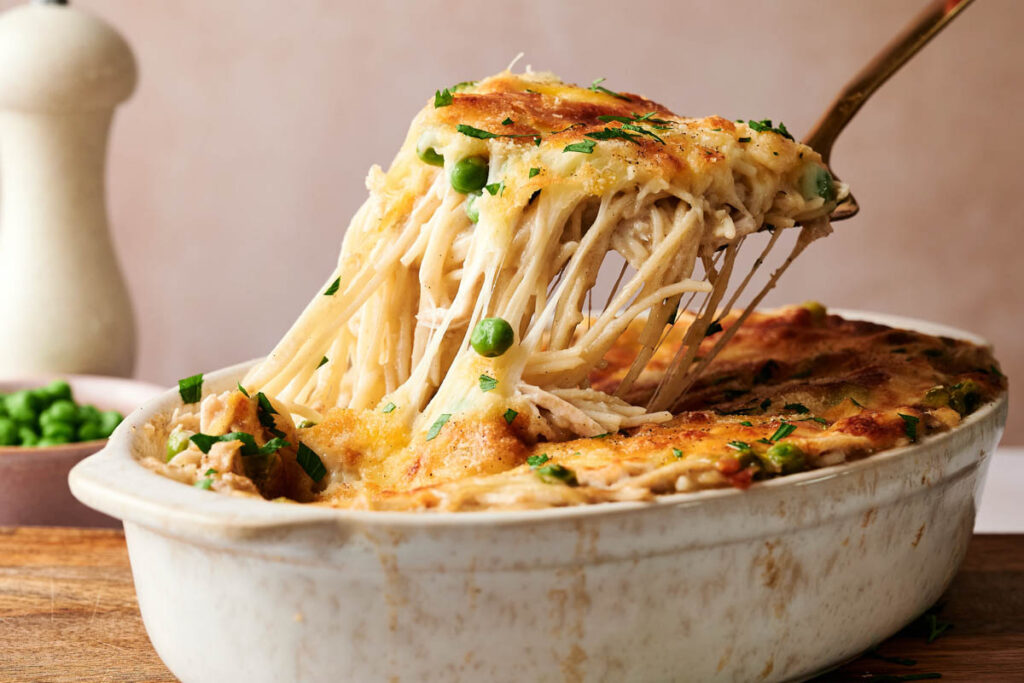 A casserole dish with a serving of Chicken Tetrazzini being lifted out, revealing baked pasta, melted cheese, peas, and a golden-brown top.