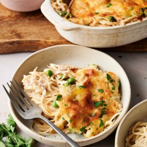 A serving of baked spaghetti with melted cheese and herbs in a bowl, reminiscent of Chicken Tetrazzini, with a fork beside it. A baking dish, fresh herbs, and a bowl of green peas are in the background.