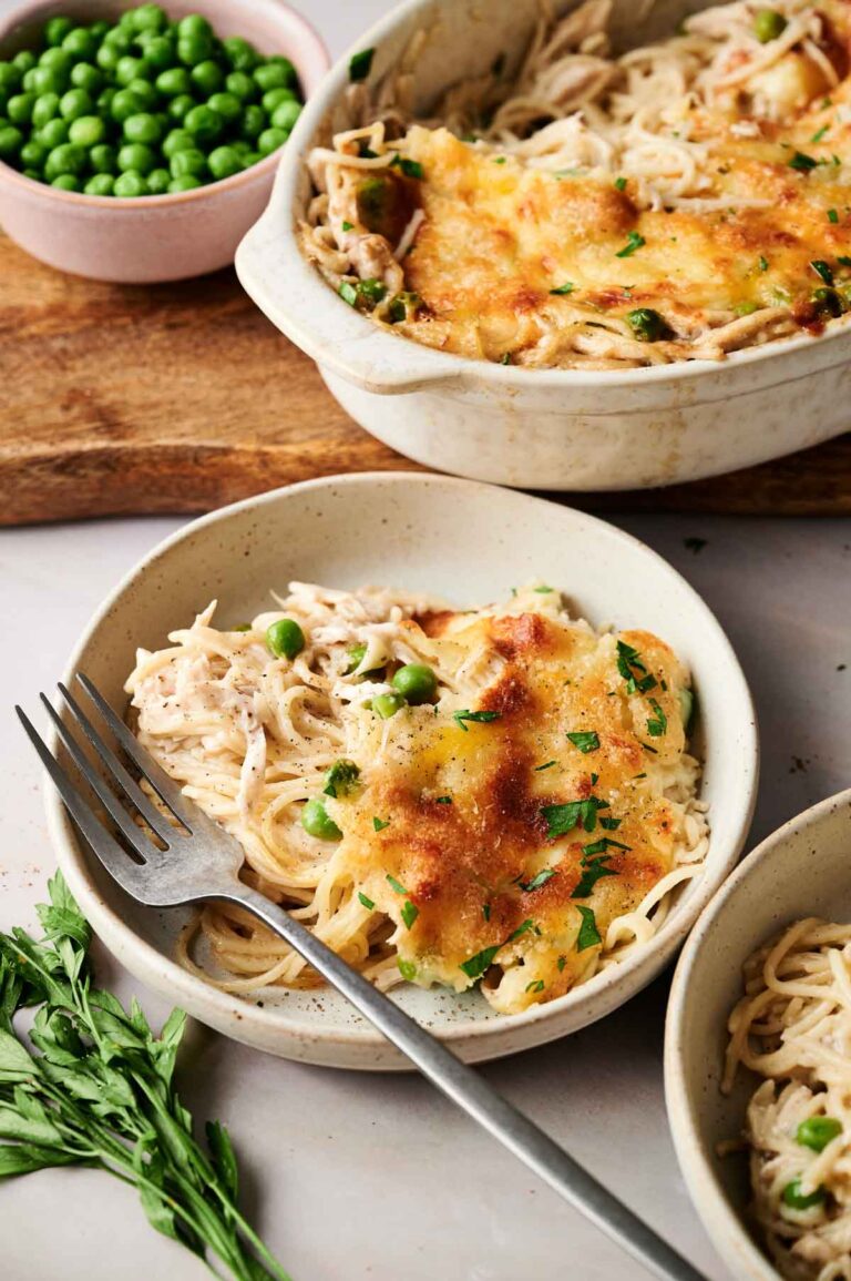 A serving of baked spaghetti with melted cheese and herbs in a bowl, reminiscent of Chicken Tetrazzini, with a fork beside it. A baking dish, fresh herbs, and a bowl of green peas are in the background.