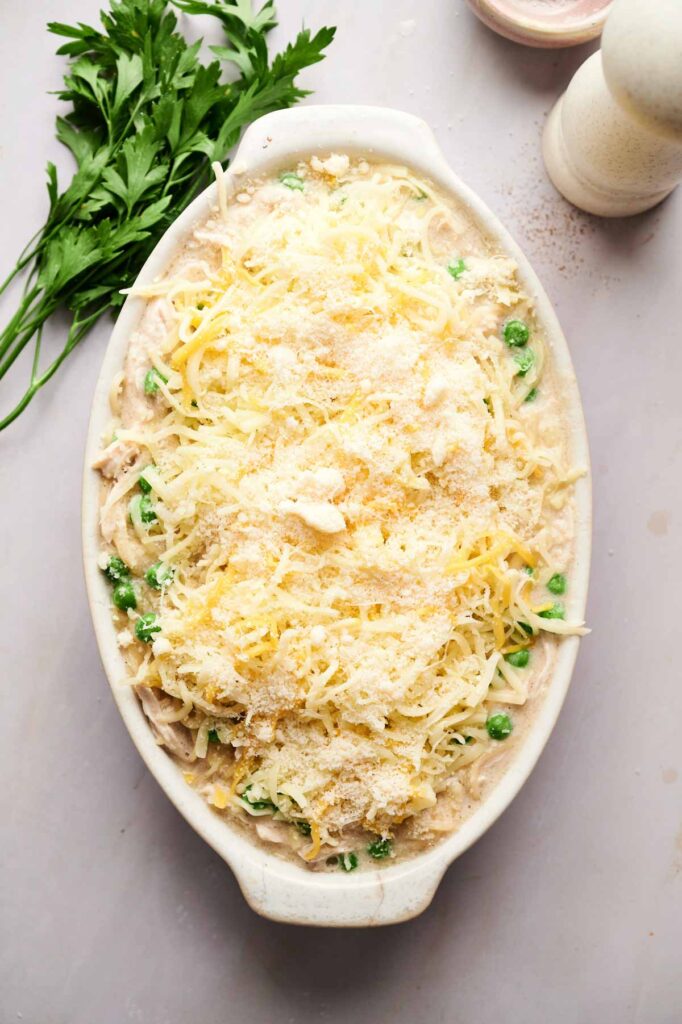 An oval baking dish filled with unbaked Chicken Tetrazzini, topped with shredded cheese, sits next to fresh parsley and a pepper grinder on a light surface.